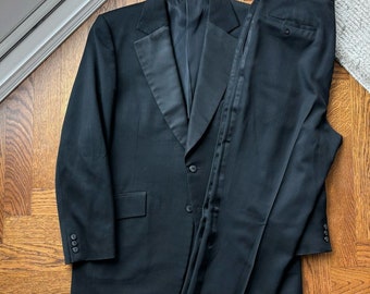 May include: A black tuxedo suit with a satin lapel. The jacket has a single button closure and the pants have a side stripe.