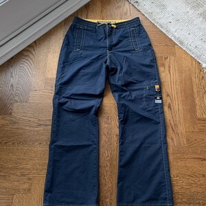 May include: Dark blue trousers with yellow stitching and a small yellow patch on the right leg. The trousers have a drawstring waist and multiple pockets.