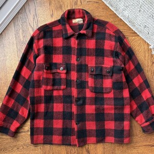 May include: A red and black checked wool shirt jacket with two chest pockets and button fastenings. The jacket features a collared neckline and long sleeves. The classic lumberjack style is visible in the bold, checkered pattern.