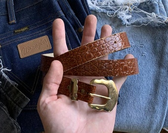 May include: A brown leather belt with a textured, crocodile-like pattern and a gold-colored buckle. The belt is held in front of a pair of blue denim jeans. The jeans have a Wrangler label.