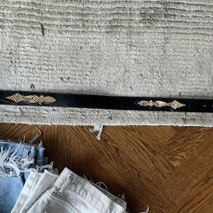 Op de afbeelding: Een zwarte leren riem met een gouden slangenhuidontwerp. De riem ligt op een lichtgrijs tapijt.