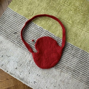 May include: A handmade red felt bag with a rounded body and a long handle. The bag is made of a thick, textured material. The bag is lying on a rug with green, gray, and white stripes.