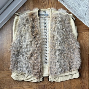 May include: A beige vest with a fur front and a patterned interior. The vest has a cream-colored leather trim and a textured, woven interior with black and white stripes. The fur is a mix of brown and gray tones.