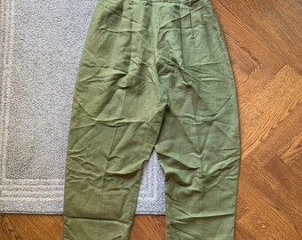 May include: Green linen pants with a high waist and pleated front. The pants are a solid olive green color and appear to be made of a lightweight fabric. The pants are folded and lying on a light gray rug and a wooden floor.
