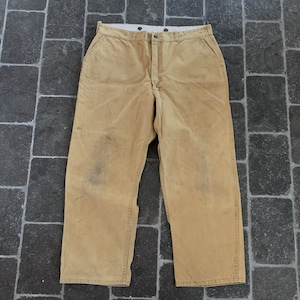 Könnte beinhalten: Eine beige Arbeitshose mit weißem Bund und Knopfverschluss. Die Hose hat einen abgenutzten Look und ist aus strapazierfähigem Stoff gefertigt.