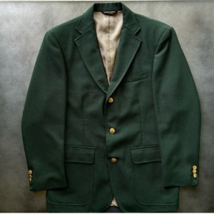 Puede incluir: Una chaqueta de sport verde oscuro con botones dorados. La chaqueta tiene un cierre de un solo botón y dos bolsillos delanteros.