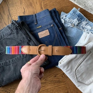 May include: A brown leather belt with a silver buckle and a colourful woven strap. The strap has red, orange, yellow, green, blue, and purple stripes.