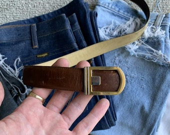 May include: A brown leather belt with a gold-colored buckle is displayed. The belt is reversible, with a tan leather side visible. The belt is shown over a pair of blue jeans.
