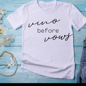Op de afbeelding: Wit T-shirt met de tekst "vino before vows" in zwarte cursieve letters.