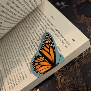 May include: A bookmark shaped like a monarch butterfly with orange, black, and white wings, positioned between the pages of an open book. The book displays printed text on white paper, with the word EMMA and the page number 105 visible.