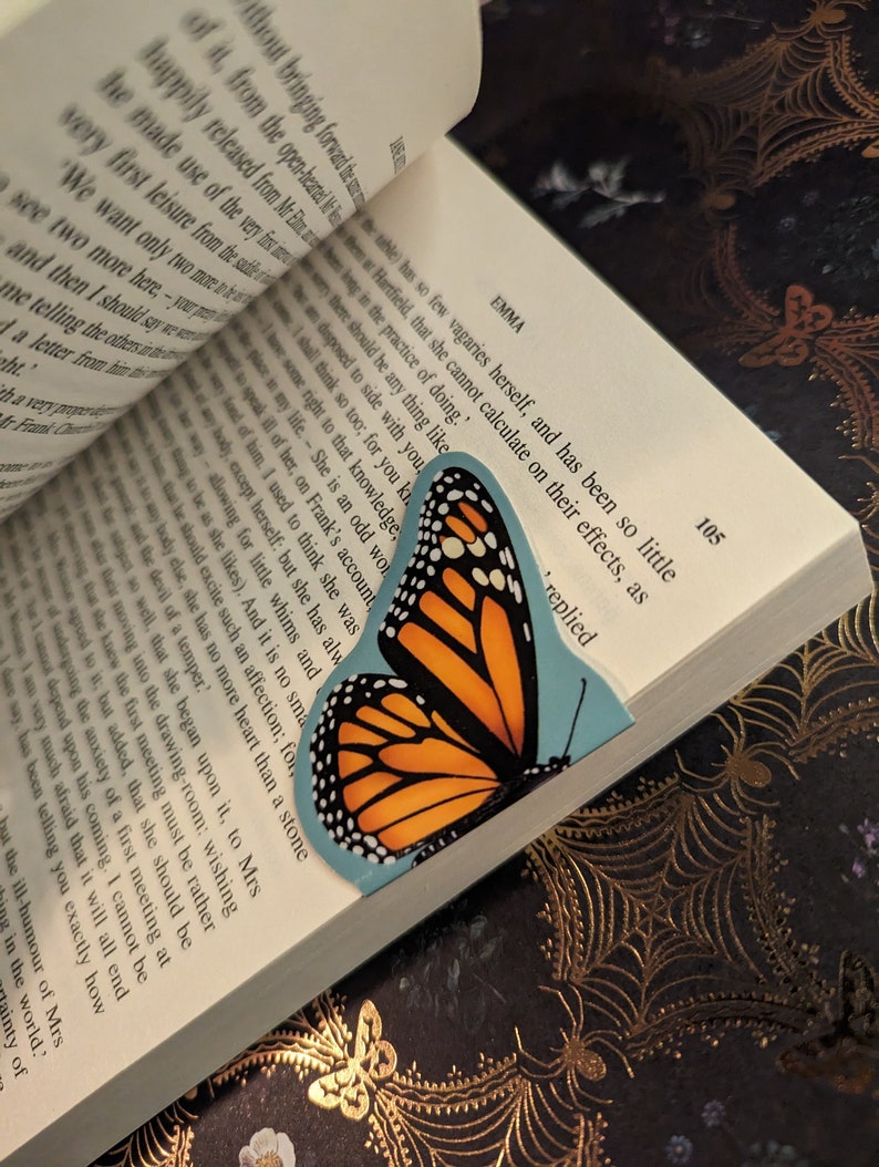 May include: A bookmark shaped like a monarch butterfly with orange, black, and white wings, positioned between the pages of an open book. The book displays printed text on white paper, with the word EMMA and the page number 105 visible.