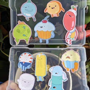 May include: A clear plastic storage case with a variety of colorful cartoon character stickers. The stickers feature characters from the animated television series Adventure Time. The stickers are in a variety of shapes and sizes, and they are all brightly colored.