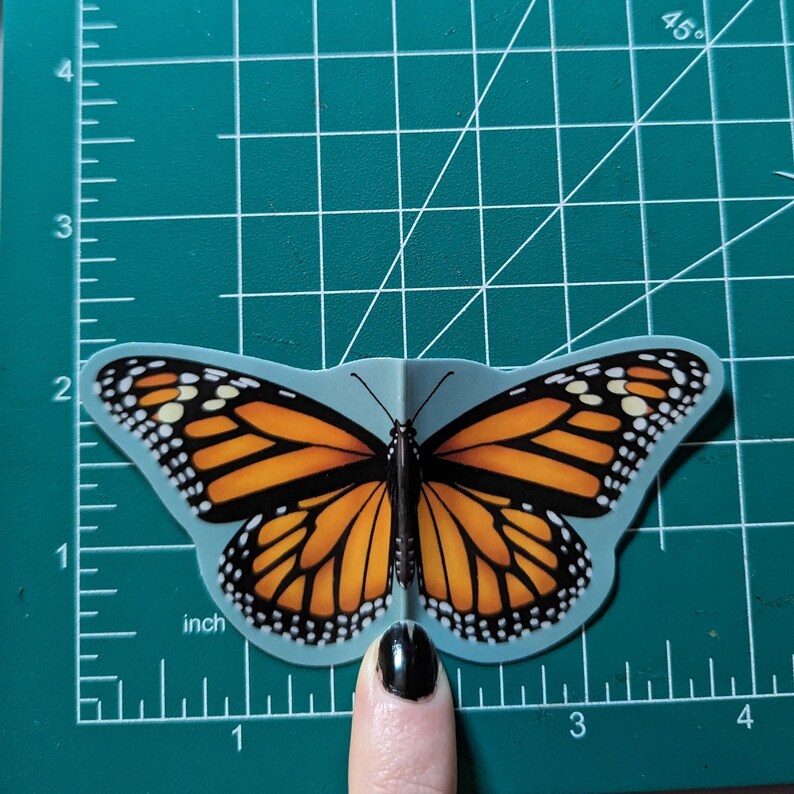 May include: A monarch butterfly sticker with orange, black, and white wings positioned on a green cutting mat with a visible ruler. The sticker measures approximately 3.5 inches in width and 2 inches in height as shown by the grid markings.