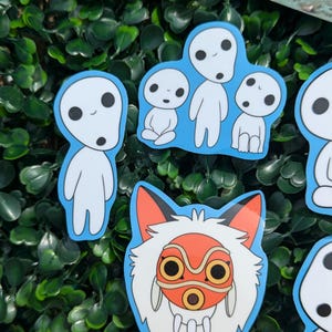 May include: A selection of stickers depicting characters from the animated film Princess Mononoke. The stickers show white Kodama spirits with black eyes and a fox-like character with a red mask and orange details, all with a blue border.