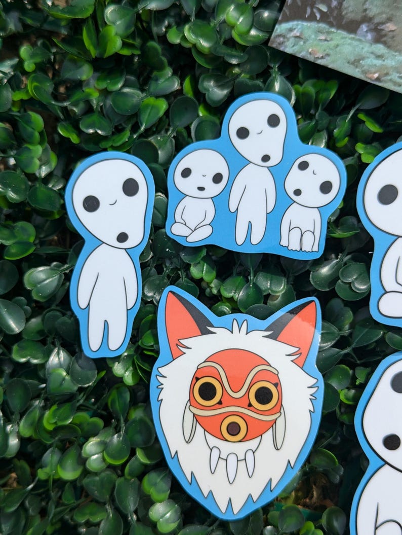 May include: A selection of stickers depicting characters from the animated film Princess Mononoke. The stickers show white Kodama spirits with black eyes and a fox-like character with a red mask and orange details, all with a blue border.