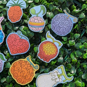 Puede incluir: Un conjunto de nueve pegatinas coloridas con diferentes dise&ntilde;os de la Fruta del Diablo del anime One Piece. Las pegatinas est&aacute;n sobre un fondo verde.