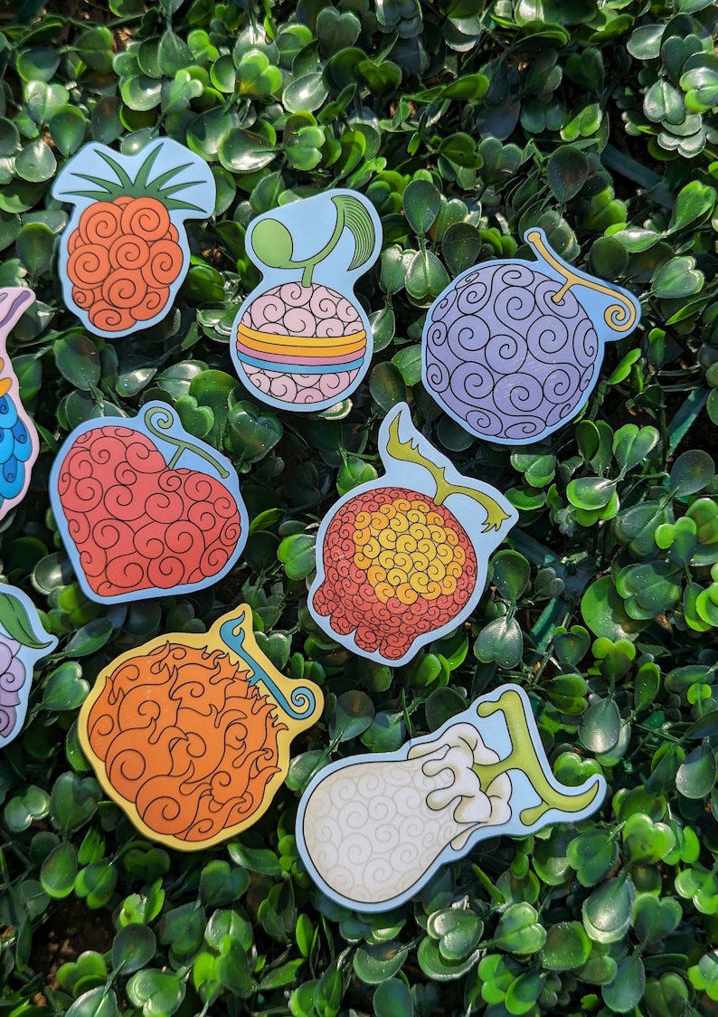 Puede incluir: Un conjunto de nueve pegatinas coloridas con diferentes dise&ntilde;os de la Fruta del Diablo del anime One Piece. Las pegatinas est&aacute;n sobre un fondo verde.