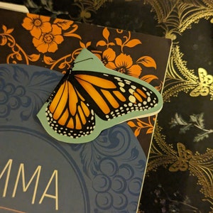 May include: A butterfly-shaped bookmark featuring a vibrant orange and black monarch butterfly wing pattern on a light blue background, resting on the cover of a book titled Emma by Jane Austen with orange floral and blue circular designs.