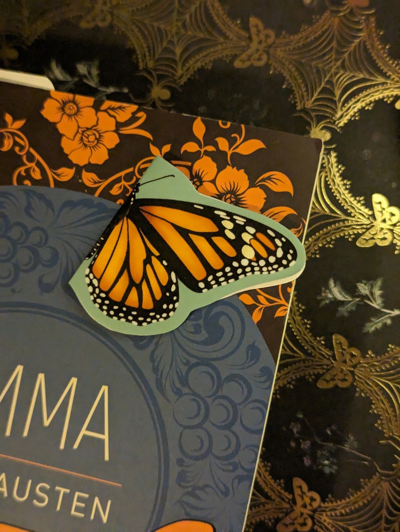 May include: A butterfly-shaped bookmark featuring a vibrant orange and black monarch butterfly wing pattern on a light blue background, resting on the cover of a book titled Emma by Jane Austen with orange floral and blue circular designs.