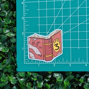 May include: A die-cut sticker of a red book with a yellow handprint and the number 3 on the cover, featuring a bookmark and abstract line art on the side, placed on a green cutting mat with white grid lines measuring approximately 2 inches wide.