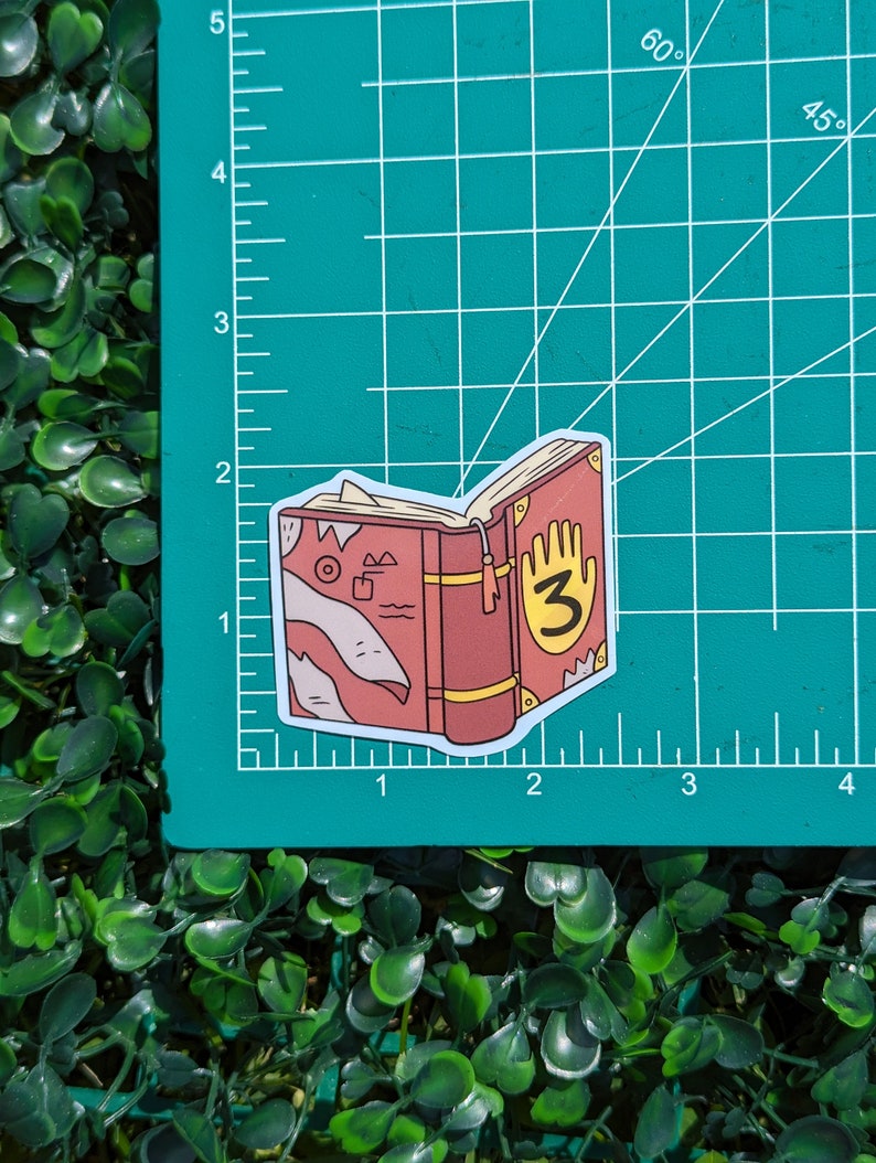 May include: A die-cut sticker of a red book with a yellow handprint and the number 3 on the cover, featuring a bookmark and abstract line art on the side, placed on a green cutting mat with white grid lines measuring approximately 2 inches wide.