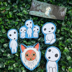 May include: A selection of stickers depicting characters from a Japanese animated film. The stickers show various poses of the forest spirits, outlined in blue, and a fox mask with red and orange details. A small print of a forest scene is also included.