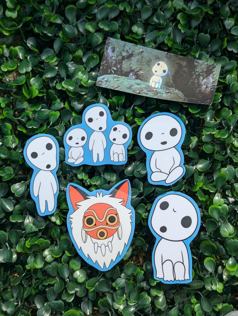 May include: A selection of stickers depicting characters from a Japanese animated film. The stickers show various poses of the forest spirits, outlined in blue, and a fox mask with red and orange details. A small print of a forest scene is also included.