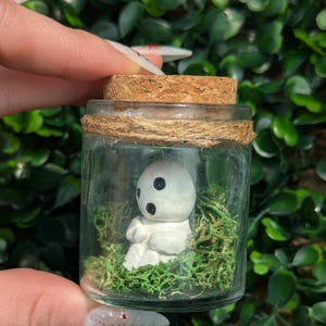 May include: A small glass jar with a cork stopper and twine detail, containing a white figurine with black eyes and a mossy base. The figurine is in a seated position. The jar is held against a backdrop of green foliage.