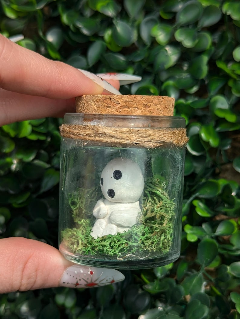 May include: A small glass jar with a cork stopper and twine detail, containing a white figurine with black eyes and a mossy base. The figurine is in a seated position. The jar is held against a backdrop of green foliage.