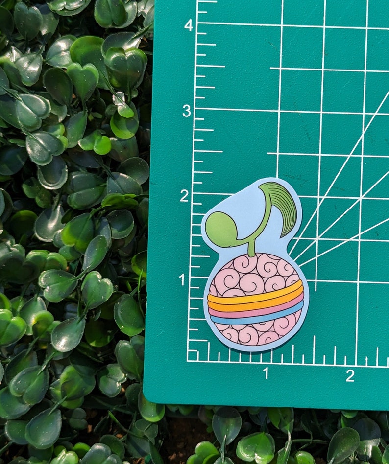 Puede incluir: Una pegatina rosa y azul de una fruta de dibujos animados con un tallo verde y tres bandas de colores alrededor del medio. La fruta est&aacute; sobre una estera de corte verde con l&iacute;neas blancas.