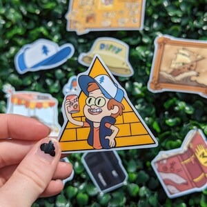 May include: A die-cut vinyl sticker featuring a cartoon character with brown hair wearing a blue and white cap, an orange shirt, and a dark vest. The character stands in front of a yellow triangle with brick patterns and holds a pink soda can.