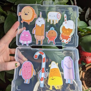 May include: A clear plastic storage case with a variety of colorful cartoon food stickers. The stickers feature characters like a donut, a candy cane, a popsicle, and a cake.