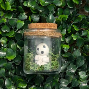 May include: A glass jar with a cork lid, bound with twine, holds a small white figurine with black features, nestled in green moss. The jar is set against a background of green leaves.