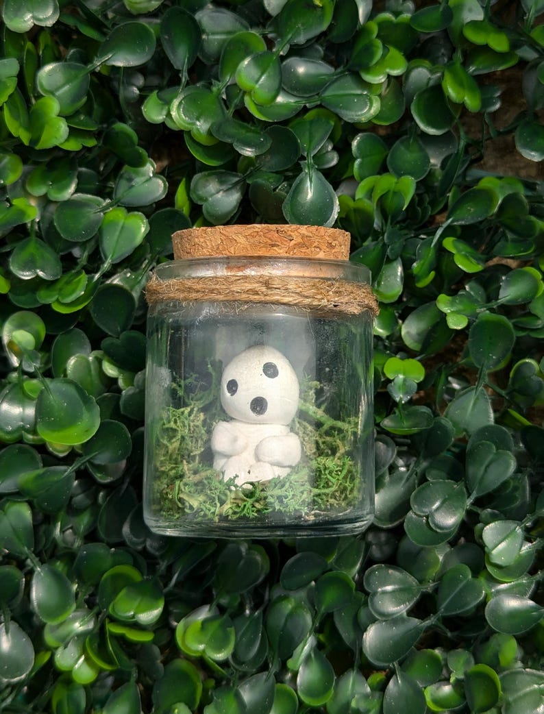 May include: A glass jar with a cork lid, bound with twine, holds a small white figurine with black features, nestled in green moss. The jar is set against a background of green leaves.