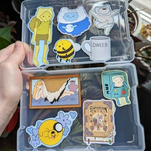May include: A clear plastic storage case filled with Adventure Time character stickers. The stickers feature Finn the Human, Jake the Dog, BMO, Ice King, Marceline the Vampire Queen, and the Enchiridion.