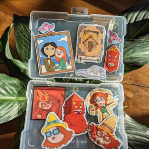 May include: A clear plastic container filled with colorful cartoon stickers. The stickers feature various characters and objects, including a book with the word "Enchiridion", a framed picture, and several character portraits. The stickers are in various shapes and sizes.