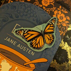 May include: A monarch butterfly sticker with orange, black, and white wings is placed on a blue book cover featuring the text Jane Austen and an orange teacup illustration. The sticker has a light blue border and is positioned over the book title.
