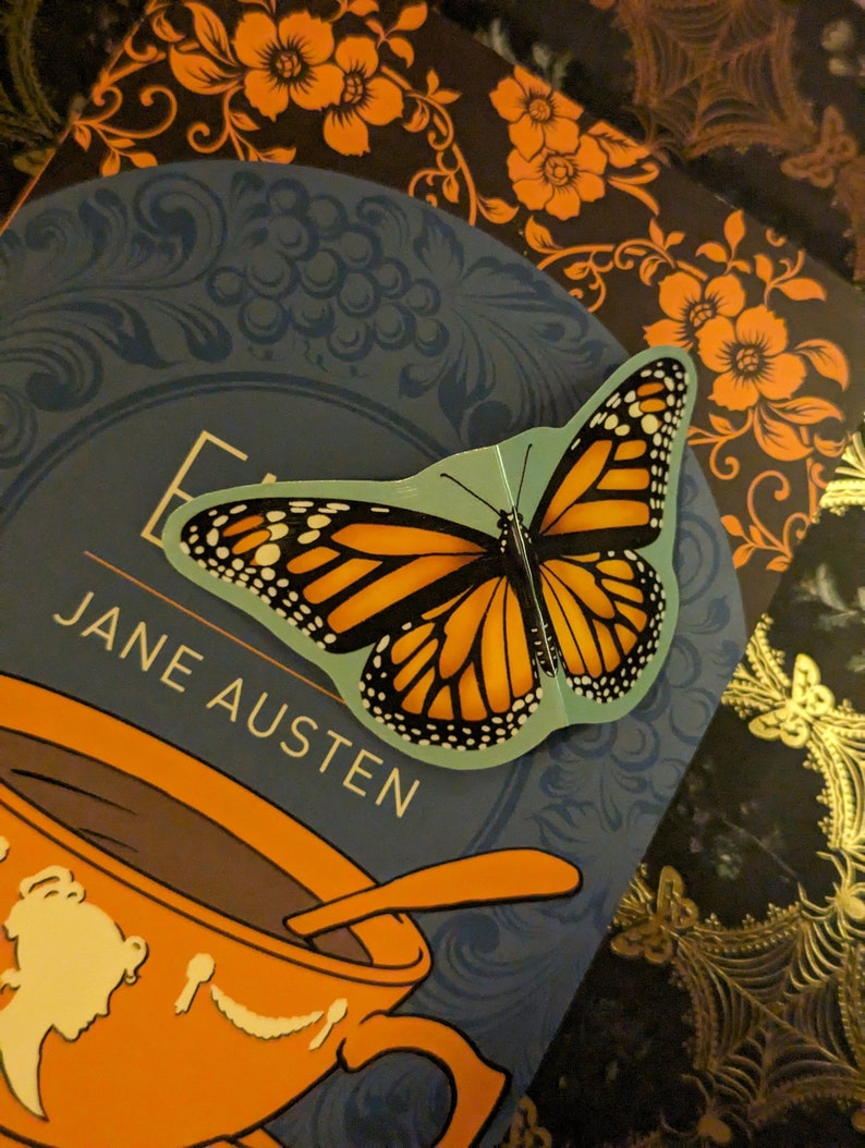 May include: A monarch butterfly sticker with orange, black, and white wings is placed on a blue book cover featuring the text Jane Austen and an orange teacup illustration. The sticker has a light blue border and is positioned over the book title.