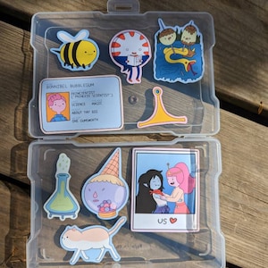 May include: A clear plastic storage case with a variety of colorful stickers featuring characters from the animated television series Adventure Time. The stickers include a bee, a candy person, a mushroom, a snake, a cat, a princess, a wizard, and a photo of two characters. The case has a label that reads "BONNIBEL BUBBLEGUM PRINCIENTIST (PRINCESS SCIENTIST) SCIENCE MAGIC ABOUT YAY BIG ONE GUMSWORTH".