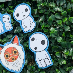 May include: A selection of stickers depicting characters from a Japanese animated film. The stickers show a white spirit with black eyes and a fox-like creature with an orange mask, all with a blue border. The stickers are on a green leafy background.