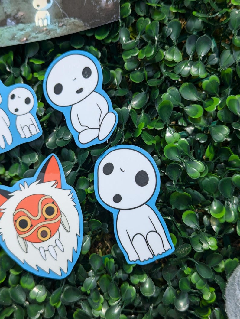 May include: A selection of stickers depicting characters from a Japanese animated film. The stickers show a white spirit with black eyes and a fox-like creature with an orange mask, all with a blue border. The stickers are on a green leafy background.