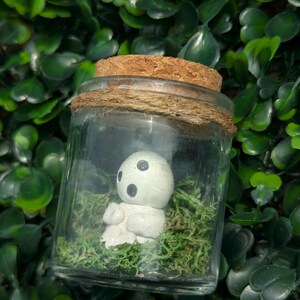 May include: A glass jar with a cork lid and twine detail, housing a small white figurine with black eyes and moss. The jar is set against a backdrop of green foliage.