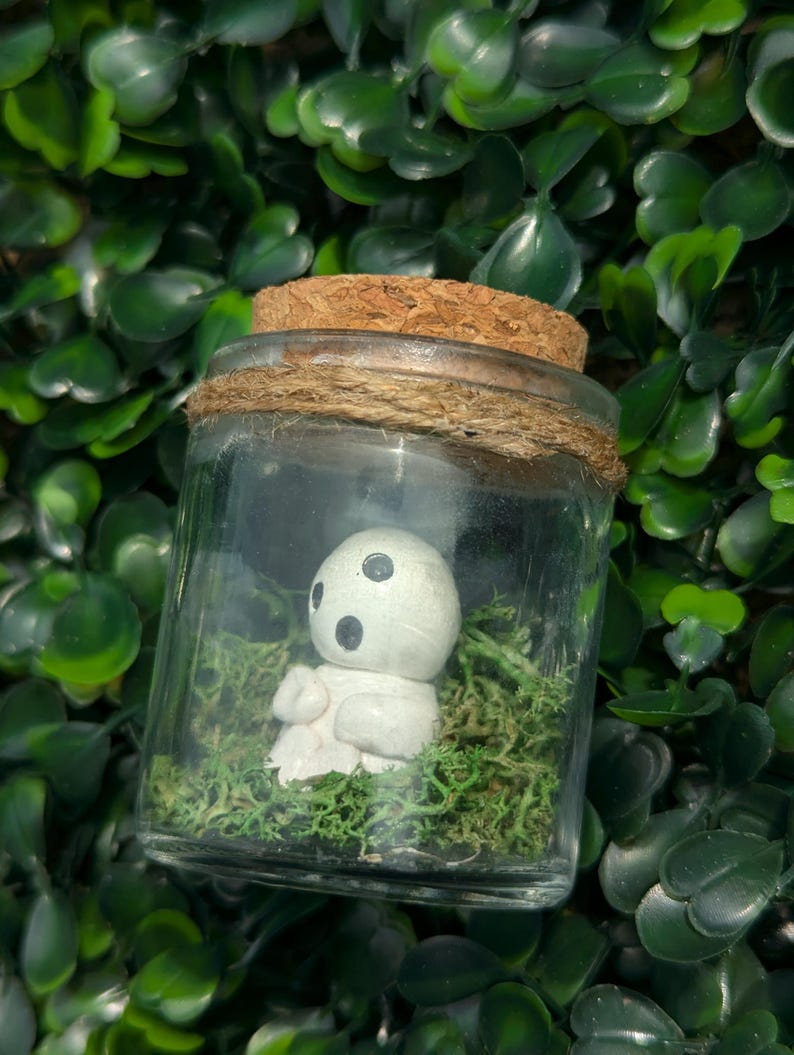May include: A glass jar with a cork lid and twine detail, housing a small white figurine with black eyes and moss. The jar is set against a backdrop of green foliage.