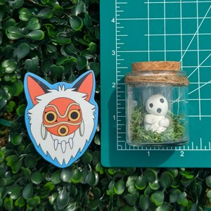 May include: A sticker depicting a wolf-like character with a red and orange mask, and a small glass jar with a cork lid containing a white figurine and green moss. The items are on a green surface.