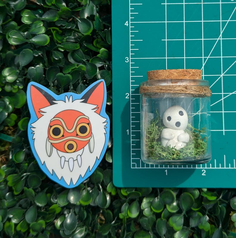 May include: A sticker depicting a wolf-like character with a red and orange mask, and a small glass jar with a cork lid containing a white figurine and green moss. The items are on a green surface.