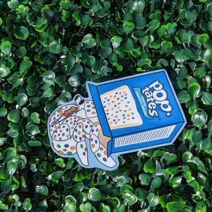 May include: A blue and white sticker of a Pop-Tarts box with a confetti cake inside. The cake is being pulled out of the box by a hand with confetti sprinkles on it. The text on the box reads "Pop-Tarts Confetti Cake"