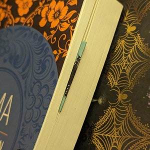 May include: A book with a dark cover featuring orange floral patterns and a blue circular design, with a thin light blue bookmark inserted between the pages. The book is positioned against a dark background decorated with gold spider web patterns.