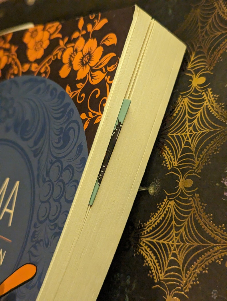 May include: A book with a dark cover featuring orange floral patterns and a blue circular design, with a thin light blue bookmark inserted between the pages. The book is positioned against a dark background decorated with gold spider web patterns.