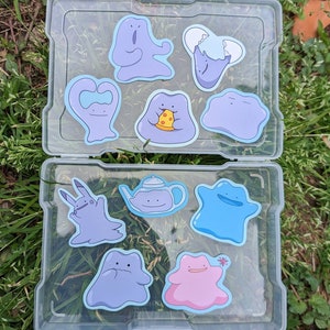 May include: A clear plastic storage case containing ten die-cut vinyl stickers featuring various purple, blue, and pink amorphous blob-like characters. The stickers depict characters in different poses, including holding a slice of pizza, wearing an eggshell, shaped like a teapot, and holding a small flower.