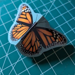 May include: A three-dimensional monarch butterfly sticker featuring vibrant orange and black wings with white spots along the edges. The sticker is folded along the center body, creating a raised effect that mimics the appearance of a butterfly in flight.